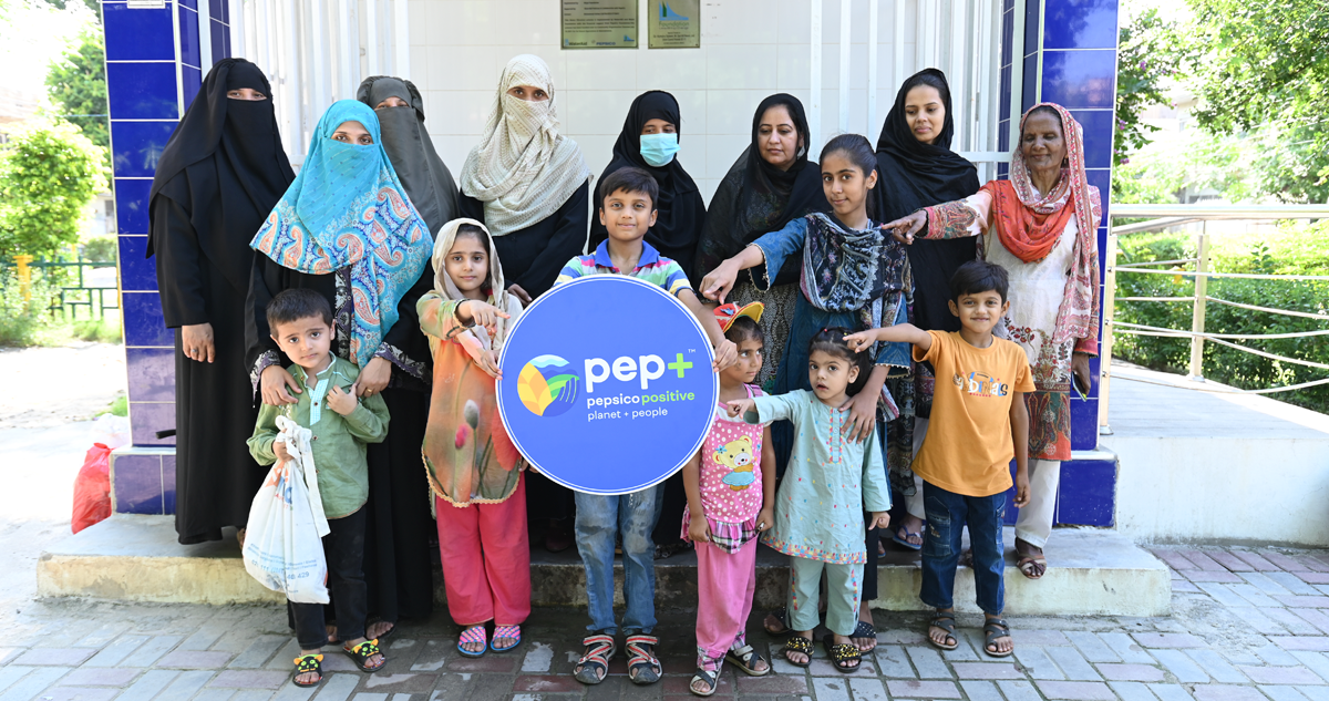 Women and children holding and pointing to pep+ PepsiCo Positive sign